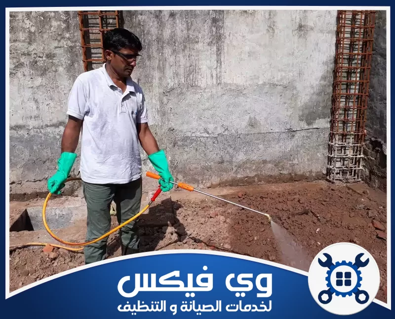 فحص تربة قبل رش الدفان في حفر الباطن 0544909308 شركة وي فيكس فني داخل موقع إنشائي يرتدي قفازات واقية ويستخدم معدات مخصصة لمعالجة التربة بجوار أساسات المبنى.